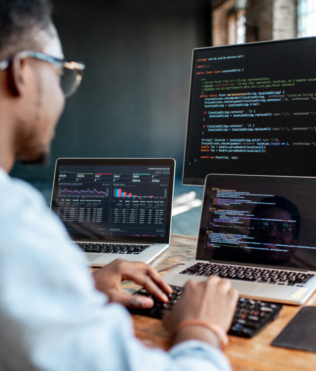 Young african male programmer writing program code sitting at the workplace with three monitors in the office. Image focused on the screen